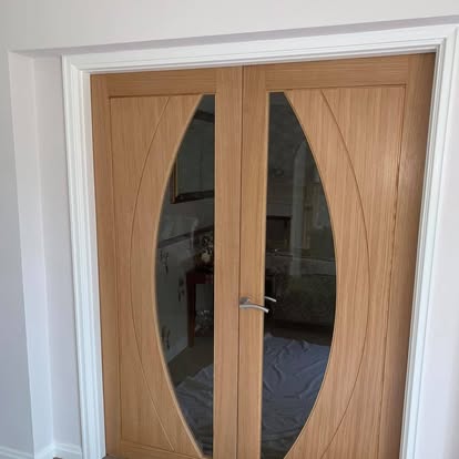 Light oak internal double doors with oval glass panels and lever handle, opening to a room with protective dust sheets on the floor.