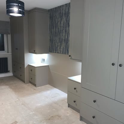 Bedroom renovation with light grey floor-to-ceiling wardrobes, central desk niche with LED strip lighting, textured blue feature wall, and unfinished subfloor.