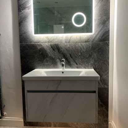Modern bathroom with wall-mounted white vanity and integrated sink, dark marble-effect wall tiles, and large backlit mirror with magnifying detail.