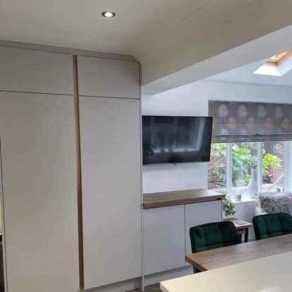 Contemporary living space with tall light-grey storage units, wood-grain TV sideboard, wall-mounted television, and green velvet dining chairs by a window.