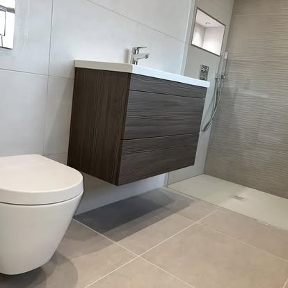 Modern bathroom with wood-grain floating vanity, wall-hung toilet, walk-in shower with textured beige feature tiles, and large-format grey wall and floor tiles.
