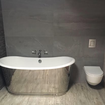 Luxury bathroom with reflective chrome freestanding tub, grey stone wall tiles, wood-effect floor planks, and wall-hung toilet with square flush plate.