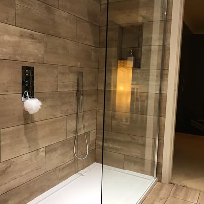 Walk-in shower with warm oak wood-effect wall tiles, chrome handheld shower on a slide rail, recessed tiled niche, and frameless glass enclosure.