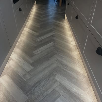 Top-down view of grey wood-effect herringbone kitchen flooring between light shaker cabinets with warm LED toe-kick lighting.
