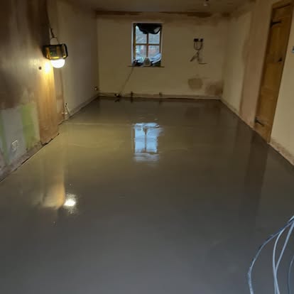 Room mid-renovation with glossy dark grey self-leveling floor coating reflecting window light, unfinished plaster walls, and temporary work light.