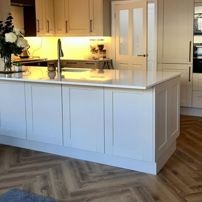 Elegant kitchen with large white shaker island, chrome faucet, herringbone wood floor, cream cabinetry with under-cabinet lighting, and floral arrangement.