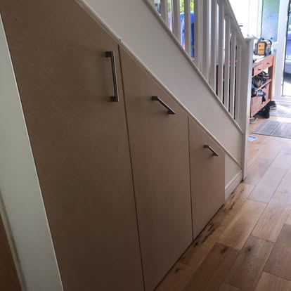 Custom fitted cabinets beneath a staircase with sloped doors and dark handles, installed over natural wood flooring in a hallway.