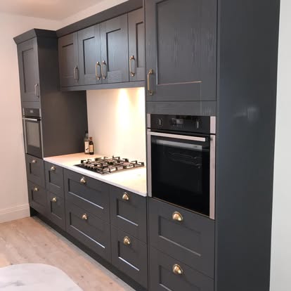 Contemporary kitchen with charcoal grey shaker cabinets, brass hardware, white countertop, integrated gas hob, and built-in black oven on light wood flooring.